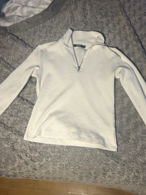 Vit ribbad tröja med halv dragkedja - Vit långärmad tröja i ribbat material med halv dragkedja framtill och hög krage. Perfekt för lager på lager och enkel att matcha med olika outfits. Tröjan har en clean och stilren look som passar till många tillfällen.