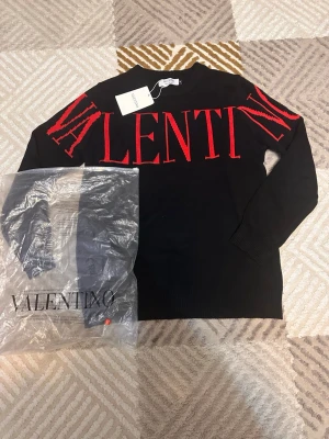 Svart stickad tröja från Valentino - Svart stickad tröja från Valentino med stor röd logotyptext över bröstet och axlarna. Tröjan har rund halsringning och långa ärmar. Tillverkad i Italien och har en klassisk, stilren design som sticker ut med den kontrasterande röda texten. Kan passa till M