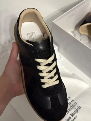 Maison Margiela🙌🤩 - Stilrena svarta sneakers från Maison Margiela med beige innersula och kontrasterande vita snören. Ovandelen är i skinn och mocka, och sulan är i gummi med klassiskt mönster. Perfekta för dig som gillar minimalistisk och lyxig design.🙌🤩