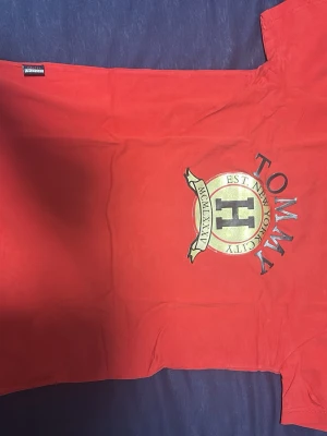 Röd Tommy Hilfiger t-shirt - Snygg röd t-shirt från Tommy Hilfiger med stort tryck i guld och svart på bröstet. Klassisk passform med korta ärmar och rund hals. Tillverkad i mjuk bomull som känns skön mot huden. Perfekt för dig som vill ha en stilren och trendig look.