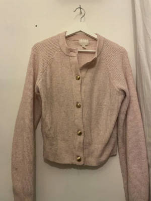 Ljusrosa stickad kofta från H&M - Säljer en ljusrosa stickad kofta från H&M med ribbad struktur och stora guldfärgade knappar framtill. Koftan har rund halsringning och långa ärmar, perfekt för lager på lager och en söt vardagslook.
