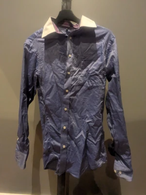 Blå skjorta från Lapious med vit krage - Snygg blå skjorta från Lapious med vit kontrastkrage och vita knappar. Insidan av kragen har ett randigt mönster i blått, vitt och rött. Skjortan är långärmad och tillverkad i bomull, perfekt för en stilren look.