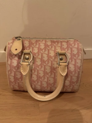  Dior Boston 20 - Säljer en  dior Boston 20  med klassiskt monogrammönster i vitt och rosa. Väskan har två korta handtag i ljust skinn, silverfärgade metalldetaljer och dragkedja upptill. Perfekt för dig som vill ha en trendig och lyxig accessoar.