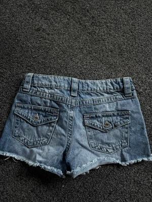 Blå jeansshorts från Gina Tricot - Säljer ett par klassiska blå jeansshorts från Gina Tricot med rå kant och två bakfickor med lock och knapp. Shortsen har bälteshällor och en avslappnad passform, perfekta för varma dagar.