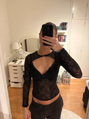 Svart spetsig långärmad topp - Säljer en svart långärmad topp i genomskinlig spets med snygg cut-out framtill. Toppen har en åtsittande passform och rund halsringning med liten knäppning. Perfekt för dig som vill ha en edgy och trendig look☺️☺️Tröjan är använd fåtal gånger!