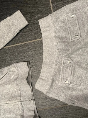 Grå mjukisbyxor med fickor - Säljer ett par grå mjukisbyxor i mjukt bomullsmaterial. Byxorna har bred resår i midjan, två fickor framtill med lock och knappdetaljer samt öljetter för eventuell snörning. Perfekta för chill dagar eller när du vill vara bekväm. Koftan är i storlek XS och byxorna i S