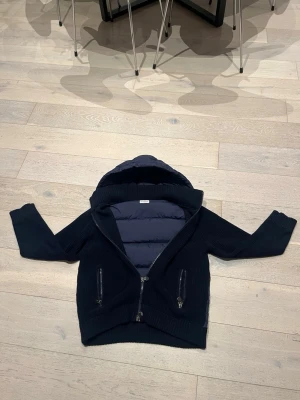 Mörkblå dunjacka med huva från moncler - Snygg mörkblå dunjacka med stickade ärmar och huva. Jackan har quiltad kropp, två dragkedjeförsedda fickor framtill och ribbade muddar. Perfekt för kyliga dagar när du vill ha både stil och värme.