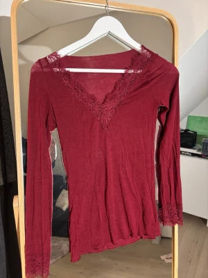Vinröd  långärmad intimissimi topp med spets  - Säljer min helt nya intimissimi tröja helt oanvänd. Snygg vinröd långärmad topp med v-ringning och spetsdetaljer vid halsringning och ärmslut. Figurnära passform och mjukt, stretchigt material som känns skönt mot huden. Perfekt för dig som vill ha en feminin och trendig look.