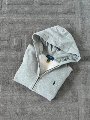 Grå hoodie från Polo Ralph Lauren - 14/16 - Snygg grå hoodie med dragkedja från Polo Ralph Lauren. Klassisk design med huva, två fickor framtill och den ikoniska lilla broderade loggan på bröstet. Perfekt för en chill och stilren look. Mjuk bomullsblandning som är skön att ha på sig.