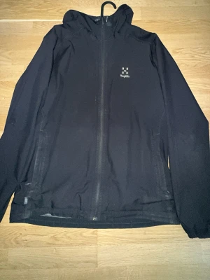 Svart vindjacka från Haglöfs - Snygg svart vindjacka från Haglöfs med huva och hel dragkedja framtill. Jackan har minimalistisk design, diskret logga på bröstet och är tillverkad i lätt syntetmaterial. Perfekt för blåsiga dagar och enkel att packa ner i väskan. Pris kan diskuteras, några skador på jackan som man kan se på bilderna luva och på höger arm 