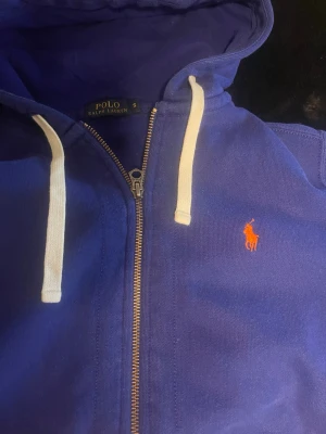 Blå hoodie från Polo Ralph Lauren - Snygg blå hoodie från Polo Ralph Lauren med dragkedja, vita snören i huvan och klassisk röd logga på bröstet. Hoodien har kängurufickor framtill och är tillverkad i mjukt bomullsmaterial. Perfekt för en avslappnad och stilren look.