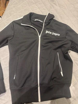 Svart track jacket från Palm Angels - Snygg svart track jacket från Palm Angels med vit dragkedja och vita ränder längs ärmarna. Jackan har hög krage, två fickor med dragkedja och logga tryckt på bröstet. Perfekt för en sportig och trendig streetwear-look.