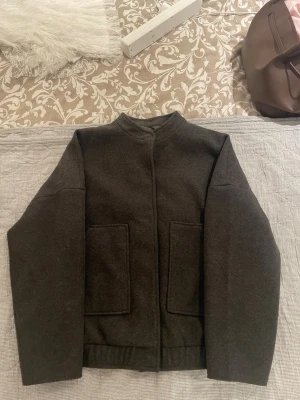 Mörkgrå ull bomberjacka - Stilren mörkgrå ”bomberjacka” i ull med raka linjer och två stora fyrkantiga fickor framtill. Jackan har en hög krage och lång ärm, perfekt för kyligare dagar. Enkel och minimalistisk design som passar till det mesta. perfekt nu till våren!