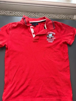 Röd pikétröja från Hilfiger Denim - Snygg röd pikétröja från Hilfiger Denim med broderad logga och text på bröstet. Tröjan har korta ärmar, klassisk krage med ränder och knappar framtill. Perfekt för en avslappnad och sportig stil.