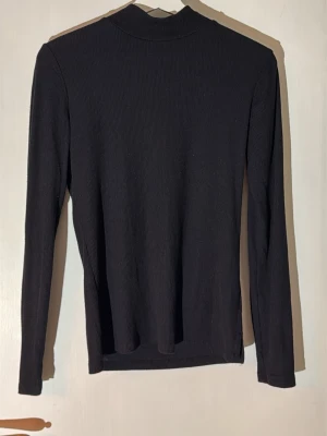 Svart ribbad långärmad tröja - En svart, ribbad långärmad tröja med hög hals. Tröjan har en smal passform och är tillverkad i ett stretchigt material som sitter snyggt på kroppen. Perfekt att styla med jeans eller kjol för en clean look.