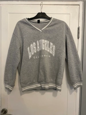 Grå sweatshirt med V-ringning - Superfin grå sweatshirt med vit text 'Los Angeles California' på bröstet. Tröjan har V-ringad hals med vit kant, ribbade muddar och nederkant med vita ränder. Perfekt för en avslappnad och sportig stil. Tyvärr för passar den inte mig längre. Lappen med butik är tyvärr avklippt så kan inte se vart den är köpt. Som ny!!