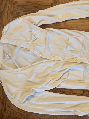 Vit kofta med fickor från Filippa K - Stilren vit kofta från Filippa K med v-ringning och knäppning framtill. Två stora bröstfickor med knappdetaljer och ribbade muddar vid ärmslut och nederkant. Perfekt att slänga över en t-shirt för en clean look.