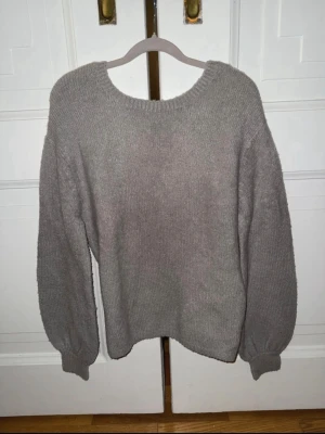 Grå oversized stickad tröja - Mysig grå stickad tröja med rund halsringning och ballongärmar, samt öppen rygg. Tröjan har en avslappnad, oversized passform och ribbade muddar vid ärmslut och nederkant. Perfekt för lager på lager och chill dagar.