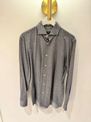Grå slim fit skjorta från Hugo Boss - Snygg grå skjorta från Hugo Boss i slim fit med små vita och mörkblå triangulära detaljer. Skjortan har klassisk krage, långa ärmar och vita knappar framtill. Perfekt för dig som gillar stilrena plagg med en twist.