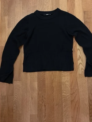 Svart stickad tröja  - En svart stickad tröja från Bik Bok med bred ribbad kant nedtill och vid ärmslut. Klassisk rund halsringning och långärmad modell. Med en slit vid ärmar och nedtill