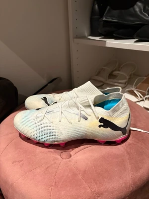 Vita Puma fotbollsskor med rosa dobbar - Säljer ett par lätta och snygga fotbollsskor från Puma i vitt med svarta loggor och rosa dobbar. Ovandelen är i syntetmaterial med snörning och en strumpliknande passform för extra support. Perfekta för dig som vill sticka ut på planen! Dom är aldrig använda, därav priset !!!!