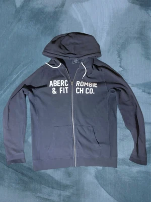 Svart hoodie från Abercrombie & Fitch - Mörkblå hoodie med dragkedja från Abercrombie & Fitch. Stor vit logga framtill, två fickor och justerbar huva med vita snören. Skön och avslappnad passform, perfekt till jeans eller joggers. Tillverkad i mjukt bomullsmaterial.