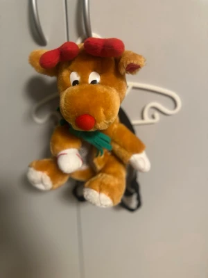 Rudolf barn ryggsäck i plysch - Söt ryggsäck formad som en ren med röda horn och nos, vita tassar och grön halsduk. Väskan är mjuk och plyschig, perfekt för dig som vill ha något gulligt och annorlunda. Svarta justerbara axelband och dragkedja på baksidan.