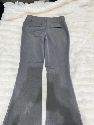 Gråa bootcut kostymbyxor - Snygga grå kostymbyxor med bootcut passform och pressveck. Byxorna har hällor för bälte och två fuskfickor bak. Perfekta för en clean och stilren look. Materialet känns som en blandning av polyester och bomull.