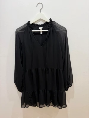 Svart volangblus från H&M - Svart blus från H&M med volangdetaljer och transparenta ärmar. Blusen har v-ringning med knytband och en lös, flowy passform. Perfekt för dig som gillar en stilren look med lite extra detaljer.