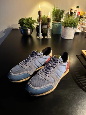 Valentino Rockrunner sneakers blå/röd - Snygga Valentino Rockrunner sneakers i blå mesh och grå mocka med röda mockadetaljer och beige skinn vid hälen. Sulan är brun med svarta nitar baktill och svart yttersula. Klassisk rund tå och snörning framtill. Perfekt för dig som gillar exklusiva sneakers med coola detaljer. Passar bra nu till kallare väder, bara höra av sig via intresse!