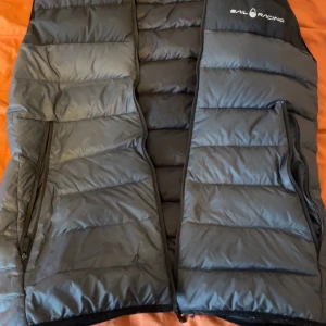 Svart dunväst från Sail Racing - Snygg svart dunväst från Sail Racing med quiltad design och två dragkedjeförsedda fickor framtill. Västen har hög krage och diskret logga på bröstet. Perfekt för lager-på-lager och håller dig varm under kyliga dagar.