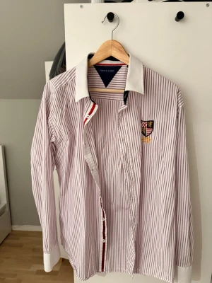 Randig skjorta Tommy Hilfiger - Säljer en vit skjorta med lila ränder från Tommy Hilfiger. Skjortan har klassisk krage och manschetter i vitt, broderad patch på bröstet och knäppning framtill. Tillverkad i bomull och har lång ärm. Perfekt för dig som gillar preppy stil.