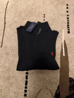 Svart ribbad polotröja Polo Ralph Lauren - Säljer en svart ribbad polotröja från Polo Ralph Lauren med half zip och röd broderad logga på bröstet. Tröjan har lång ärm och hög krage, perfekt för dig som gillar klassisk men ändå sportig stil.