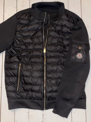 Svart cardigan jacka från Moncler (prutbart) - Svart cardigan jacka från Moncler med stickade ärmar och ribbade muddar. Jackan har dragkedja framtill, en ficka med dragkedja och Moncler-logga på ärmen. Materialmix av ull som ger både värme och stil.