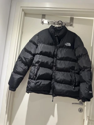 Svart pufferjacka från The North Face - Säljer en svart pufferjacka från The North Face med klassisk logga på bröstet. Jackan är quiltad, har lång ärm och dragkedja framtill. Perfekt för kalla dagar med sin tjocka och fluffiga design. 700-märkning på ärmen.