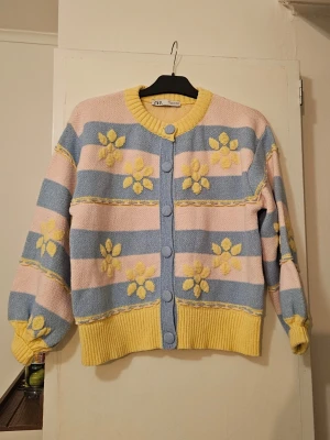 Pastellrandig kofta med blommor Zara - Söt kofta från Zara i pastellfärger med breda ränder i ljusblått och ljusrosa samt gula detaljer. Koftan har stora gula blommor, ribbstickade muddar och knappar framtill. Perfekt för dig som gillar färgglada och lekfulla plagg.