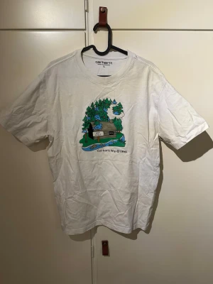 Vit Carhartt WIP t-shirt med tryck - Vit t-shirt från Carhartt WIP med ett färgglatt tryck av en stuga i skogen på bröstet. Klassisk rund hals och korta ärmar. Tillverkad i mjuk bomull som känns skön mot huden. Perfekt för dig som gillar streetwear och snygga prints.