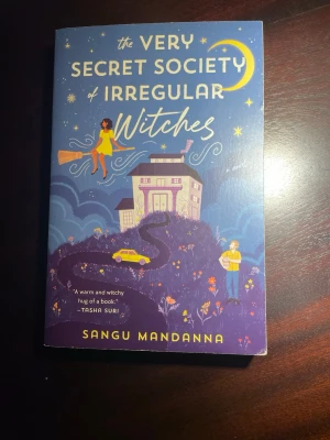 The Very Secret Society of Irregular Witches - En färgstark och magisk roman av Sangu Mandanna. Omslaget är blått med illustrationer av en häxa på kvast, ett hus, en gul bil och stjärnor. Boken har mjukt omslag och är på engelska. Perfekt för dig som gillar mysig fantasy med magi och vänskap.