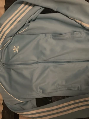 Ljusblå Adidas ziptröja - Säljer en ljusblå ziptröja från Adidas med klassiska vita ränder längs ärmarna och vit Adidas-logga på bröstet. Tröjan har hel dragkedja, ståkrage och fickor framtill. Tillverkad i ett glansigt syntetmaterial som ger en sportig look.