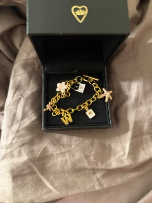 Guldigt charmarmband med berlocker - Snyggt guldigt armband med flera berlocker i olika former, bland annat en blomma, stjärna, bokstaven W och små plattor. Berlockerna har detaljer i rosa och vitt. Länken är grov och armbandet stängs med ett T-lås. Perfekt accessoar för dig som gillar personliga smycken.