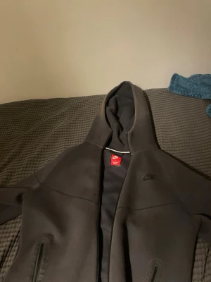 Mörk grå Nike tech hoodie med dragkedja - Snygg mörk grå hoodie från Nike med dragkedja framtill och två fickor med dragkedja. Klassisk Nike-logga på bröstet och huva med snörning. Tillverkad i mjukt och bekvämt material som passar perfekt till streetwear-looken.