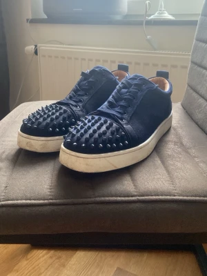 Mörkblå sneakers från Christian Louboutin - Säljer ett par mörkblå sneakers från Christian Louboutin med coola nitar på tån och snörning. Skorna har en vit platt sula och är gjorda i mocka med skinnfoder. Kommer med originalkartong och dustbag. Perfekt för dig som vill sticka ut med din stil.