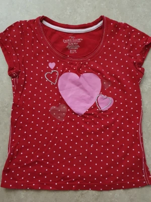 T-shirt - Söt röd t-shirt från Faded Glory med vita prickar och flera hjärtan i rosa och silver på framsidan. T-shirten har rund halsringning och korta ärmar. Perfekt för dig som gillar färgglada och lekfulla plagg.