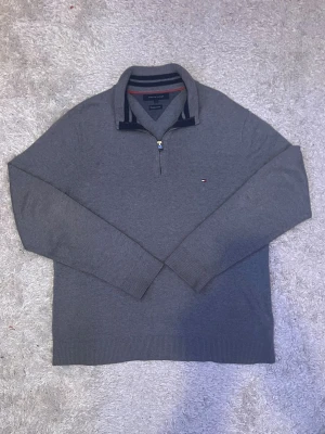 Grå half zip tröja Tommy Hilfiger - Säljer en stilren grå tröja från Tommy Hilfiger i premium bomull. Tröjan har half zip, ribbade muddar och en klassisk ståkrage med svarta och vita ränder. Liten broderad logga på bröstet. Perfekt till lager på lager under höst och vinter.