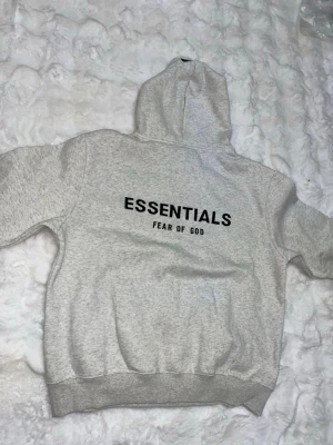 Grå hoodie Essentials Fear of God - Snygg gråmelerad hoodie från Essentials Fear of God med svart logga tryckt både fram och bak. Klassisk passform med huva och känguruficka. Tillverkad i mjukt bomullsmaterial som känns skönt mot huden. Perfekt för en avslappnad och trendig streetwear-look.