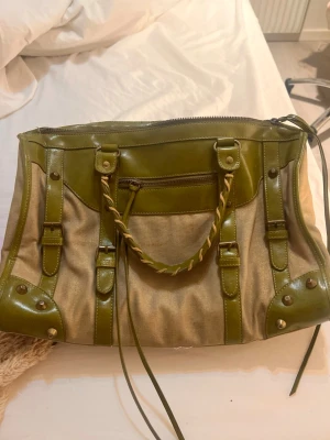 Grön och beige handväska med flätat handtag - Cool handväska i beige tyg med gröna detaljer i skinn och flätat handtag. Väskan har dragkedja upptill, dekorativa spännen och nitar samt en ficka framtill. Perfekt för dig som vill sticka ut med en unik accessoar.