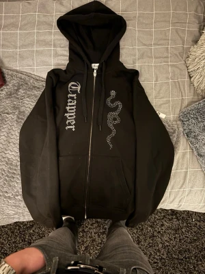 Svart zip hoodie med tryck - Säljer en svart hoodie med dragkedja från Trapper. På ena sidan finns ett coolt tryck med texten 'Trapper' i gotisk stil och på andra sidan en orm i strass. Hoodien har huva med snören och två stora fickor framtill. Perfekt för dig som gillar streetwear och vill sticka ut. Helt oanvänd.