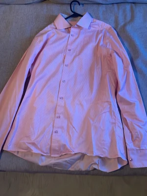 Rosa randig skjorta Oscar of Sweden - Snygg rosa och vit randig skjorta från Oscar of Sweden i slim fit-modell. Skjortan är non iron och har klassiska knappar framtill. Perfekt för dig som gillar stilrena och fräscha skjortor med modern passform. Liten defekt vid knappen, men den märker man inte av