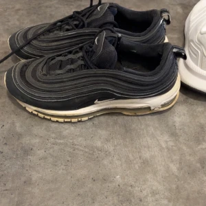 Svarta Nike Air Max 97 sneakers - Svarta Nike Air Max 97 sneakers med vågformade detaljer och vit sula. Klassisk design med snörning och diskret Swoosh-logga på sidan och tungan. Ovandel i syntet och textil, platt sula och rund tå. Perfekta för en sportig och trendig stil.