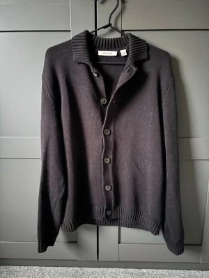 Svart kofta från Only & Sons - Otroligt schysst svart kofta från Only & Sons med knäppning framtill och bred ribbad krage. Koftan har långa ärmar och ribbade muddar vid ärmslut och nederkant. Perfekt för lager-på-lager och enkel att matcha med olika outfits. Detta plagg e bara använd ett fåtal gånger och är som helt ny! Nypris:699kr